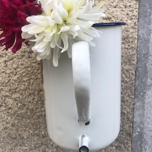 May include: A white enamelware pitcher with a blue rim and handle, filled with burgundy and white artificial flowers. The pitcher has a spout and is mounted on a textured, light brown wall.