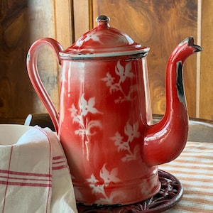 Peut inclure: Une cafetière vintage en émail rouge avec des motifs floraux blancs. La cafetière a un long bec, une poignée incurvée et un couvercle assorti. Elle repose sur un dessous de plat rouge foncé, avec un tissu à rayures rouges et blanches à côté. Un article de cuisine charmant.