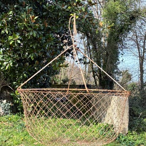 May include: A large, hanging, open-weave basket made of metal wire. The basket is suspended from a triangular frame and rope, with a rounded bottom. The basket is a rusty brown color and is hanging outdoors in front of green foliage.