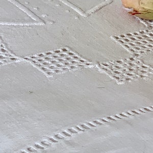 May include: Close-up of white embroidered fabric with geometric and floral patterns. The fabric features detailed stitching, including diamond and rectangular shapes, and a decorative border. A dried rose adds a touch of colour.