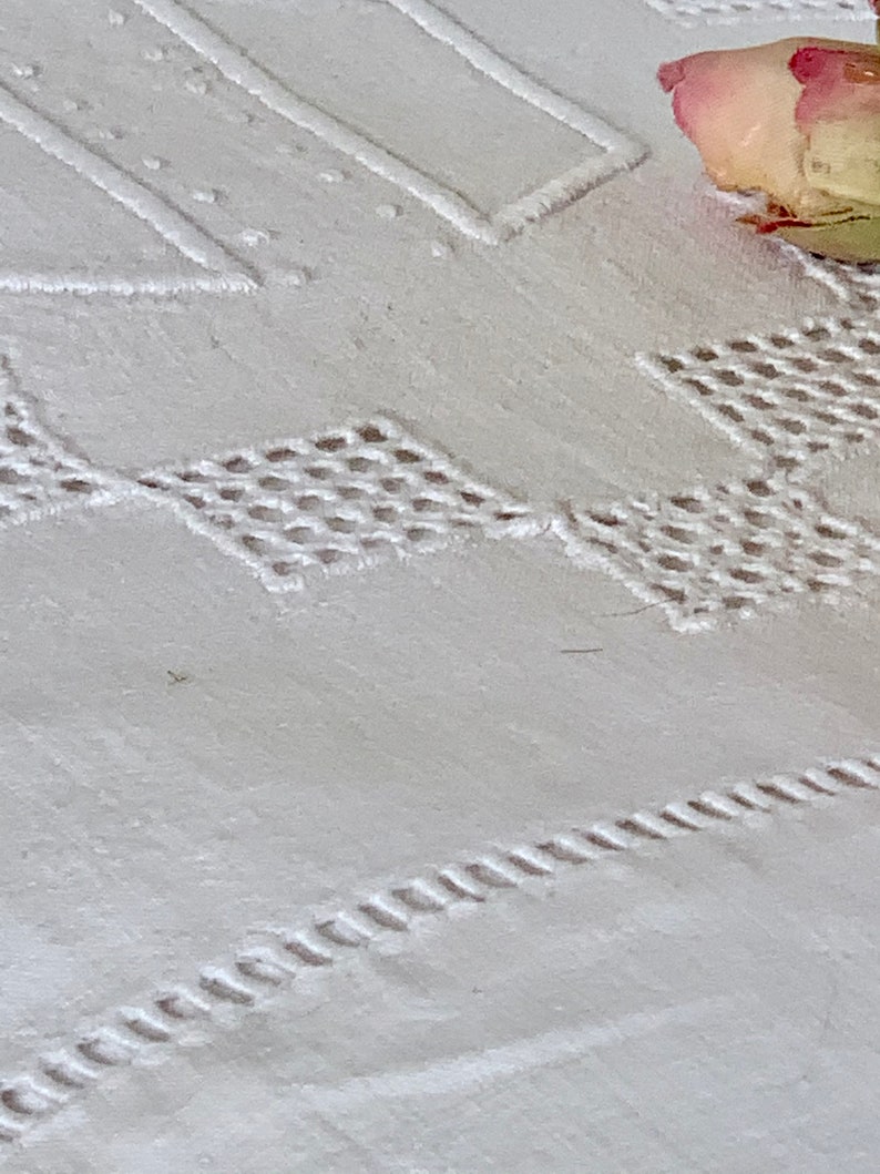 May include: Close-up of white embroidered fabric with geometric and floral patterns. The fabric features detailed stitching, including diamond and rectangular shapes, and a decorative border. A dried rose adds a touch of colour.
