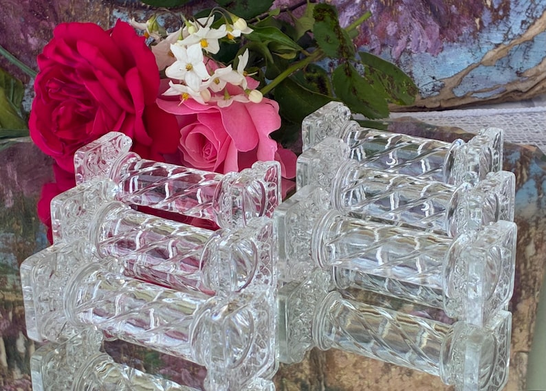 May include: Clear glass napkin rings with a diamond pattern are stacked in the foreground. The background features vibrant pink roses and white flowers. The napkin rings are reflecting the flowers and the background.