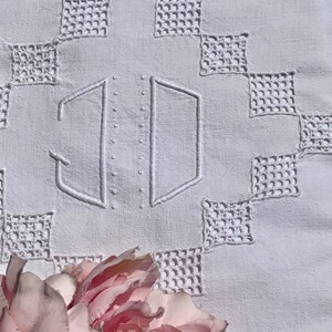 May include: A close-up of a white embroidered textile with a geometric pattern. The textile features the embroidered initials "JD" in the centre. A pink and red peony flower is in the foreground, adding a touch of colour.