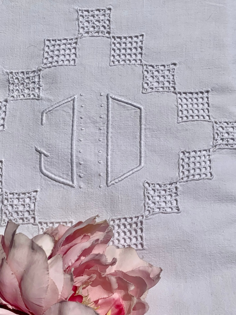May include: A close-up of a white embroidered textile with a geometric pattern. The textile features the embroidered initials "JD" in the centre. A pink and red peony flower is in the foreground, adding a touch of colour.