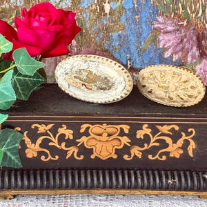 May include: A rectangular dark brown wooden box with gold-colored floral inlay, topped with two oval-shaped off-white decorative knobs. One knob features a distressed finish, while the other displays a raised gold-accented floral pattern.