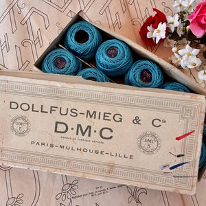 May include: A vintage cardboard box labeled DOLLFUS-MIEG & Cie D.M.C PARIS-MULHOUSE-LILLE containing multiple round balls of teal cotton embroidery thread. Each thread ball features a small circular label with the D.M.C logo and number 5.