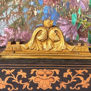 May include: A rectangular black wooden box featuring a gold-colored ornate metal crest on top. The box is decorated with light brown floral scrollwork patterns on the front panel, while the metal crest displays detailed acanthus leaf carvings.