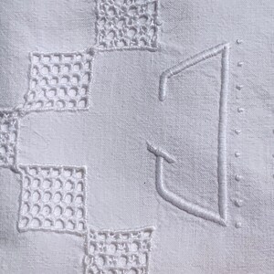 May include: Close-up of a white linen textile with embroidered details. The design features openwork squares and a large, stylized letter "J" in the centre. The embroidery is in a matching white thread, creating a subtle, elegant pattern.