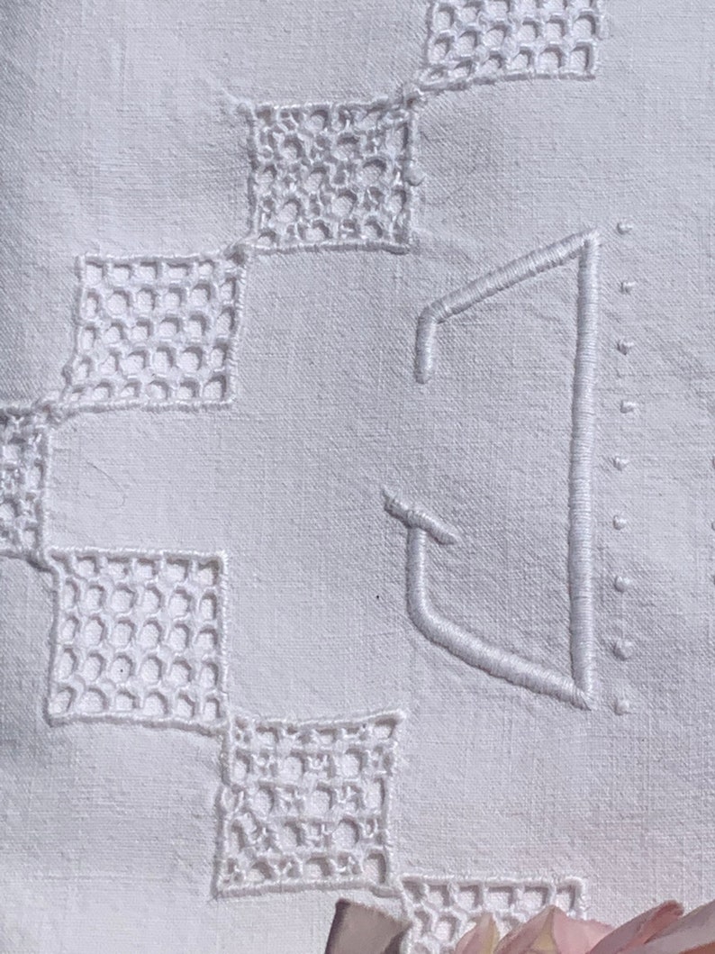 May include: Close-up of a white linen textile with embroidered details. The design features openwork squares and a large, stylized letter "J" in the centre. The embroidery is in a matching white thread, creating a subtle, elegant pattern.