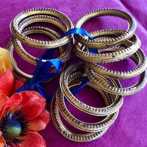 May include: Several gold-colored, oval-shaped metal frames are stacked and tied with a blue ribbon. The frames have a textured, beaded edge. Artificial orange and yellow flowers are in the lower left corner, set against a purple background.