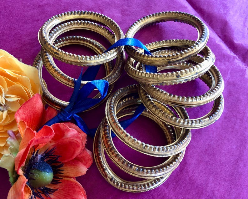May include: Several gold-colored, oval-shaped metal frames are stacked and tied with a blue ribbon. The frames have a textured, beaded edge. Artificial orange and yellow flowers are in the lower left corner, set against a purple background.