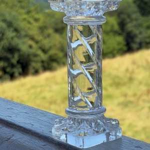 May include: A clear, glass decorative pillar with a square base and top. The pillar has a twisted, spiral design in the center. The background features a blurred view of green trees and a field.