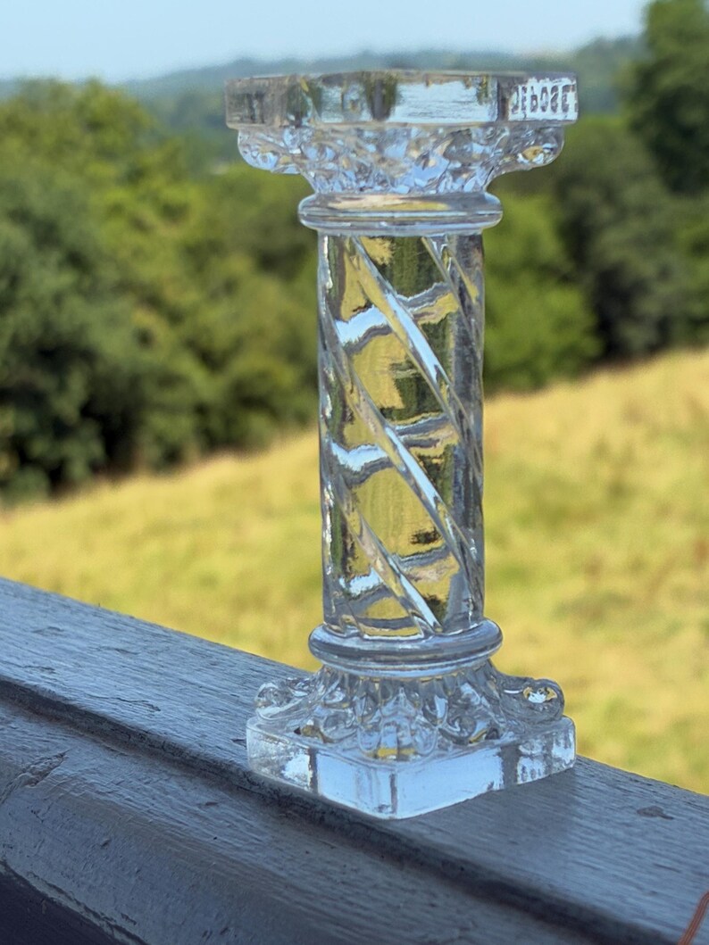 May include: A clear, glass decorative pillar with a square base and top. The pillar has a twisted, spiral design in the center. The background features a blurred view of green trees and a field.