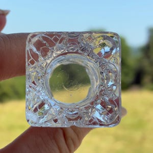 May include: A clear, square glass object with a circular indentation. The glass has a textured, patterned design on the sides. The object is held up against a blurred green and yellow background.