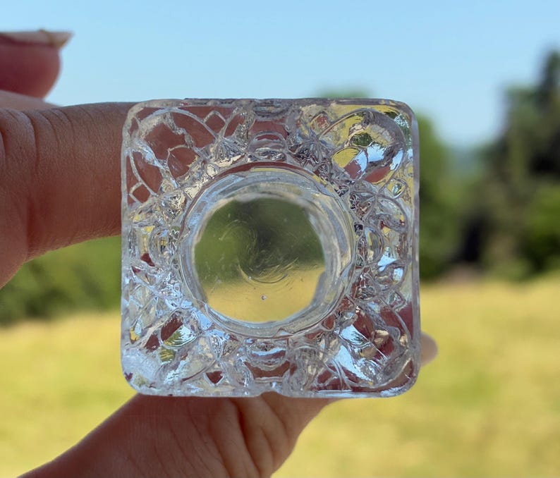 May include: A clear, square glass object with a circular indentation. The glass has a textured, patterned design on the sides. The object is held up against a blurred green and yellow background.