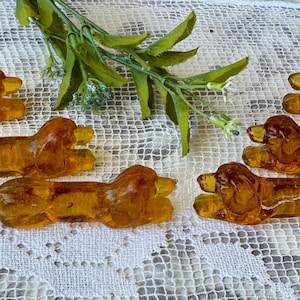 Puede incluir: Seis figuras de perro salchicha de cristal color ámbar dispuestas sobre una superficie de encaje blanco. Las figuras de perro están tumbadas con las patas extendidas. Hay follaje verde en el fondo.
