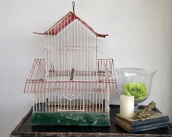 May include: A white and red birdcage with a green base. The cage has a red roof and a white wire frame. The cage is sitting on a wooden table.