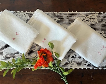 May include: Three white linen napkins with embroidered initials in red thread, arranged on a lace runner. A vibrant orange poppy and greenery add a pop of color. The napkins are folded and appear to be vintage.