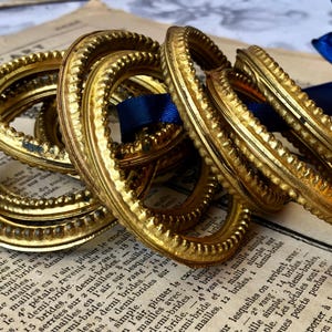 May include: Several ornate, gold-colored metal rings with a textured design are stacked on top of each other. A blue ribbon and a piece of paper with French text are in the background. The rings have a vintage aesthetic.