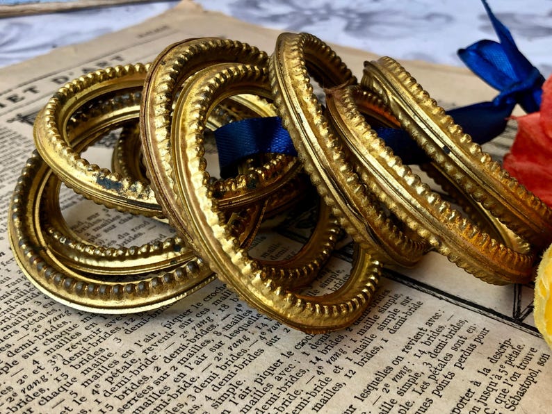 May include: Several ornate, gold-colored metal rings with a textured design are stacked on top of each other. A blue ribbon and a piece of paper with French text are in the background. The rings have a vintage aesthetic.