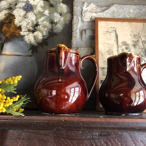 Puede incluir: Dos jarras de cerámica esmaltada marrón con un aspecto rústico. Las jarras tienen un cuerpo redondeado y un asa. Están sentadas en un estante de madera.