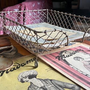 Pode incluir: Uma bandeja de mesa retangular de malha de arame com acabamento metálico, apresentando um padrão de grade e bordas curvas. A bandeja é elevada sobre quatro pequenos pés de metal e contém vários documentos de papel vintage com ilustrações.