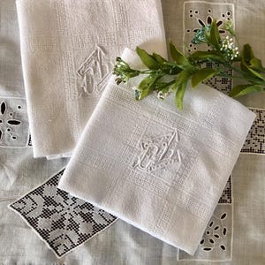 May include: Two white linen handkerchiefs with embroidered initials. The handkerchiefs are folded and placed on a white lace tablecloth.