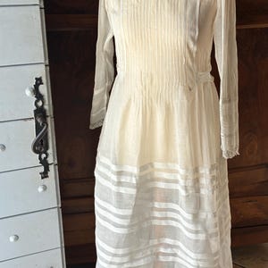 May include: A white vintage dress with a pleated bodice and a sheer overlay. The dress has a round neckline and long sleeves with lace trim. The skirt has horizontal stripes of white fabric.