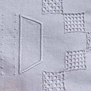 May include: Close-up of a white linen textile with embroidered geometric patterns. The design includes a large, stylized letter and openwork squares. The fabric has a subtle texture, and the embroidery is in a matching white thread.