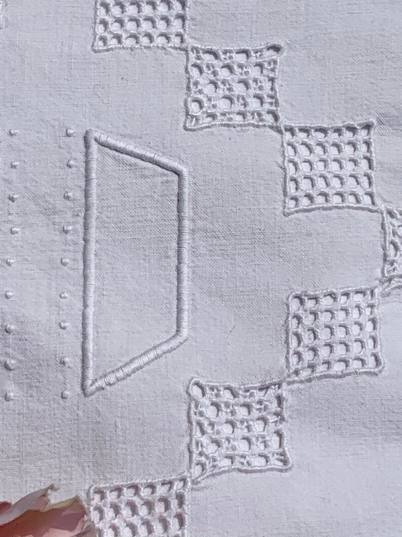 May include: Close-up of a white linen textile with embroidered geometric patterns. The design includes a large, stylized letter and openwork squares. The fabric has a subtle texture, and the embroidery is in a matching white thread.