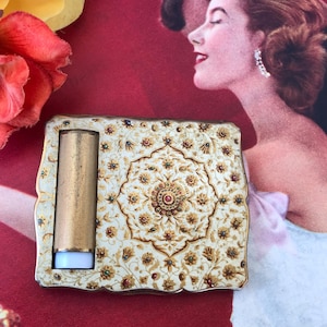 Può includere: Un compatto vintage dorato con un design floreale bianco e oro. Il compatto ha un coperchio incernierato e uno specchio rotondo piccolo all'interno. Il compatto è decorato con piccoli gioielli colorati.