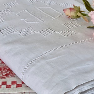 May include: White linen textile with embroidered geometric diamond patterns and the initials "JD". The folded textile has a decorative stitched border. A pink rose and green leaves are in the background. The textile is folded.
