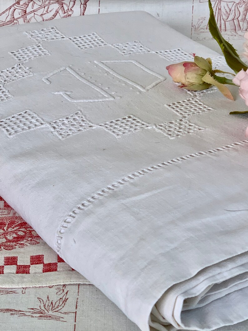 May include: White linen textile with embroidered geometric diamond patterns and the initials "JD". The folded textile has a decorative stitched border. A pink rose and green leaves are in the background. The textile is folded.