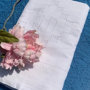May include: White linen sheet with embroidered geometric pattern and monogram. A pink peony and a silver decorative element are placed on the sheet. The sheet is folded and placed on a blue patterned surface.