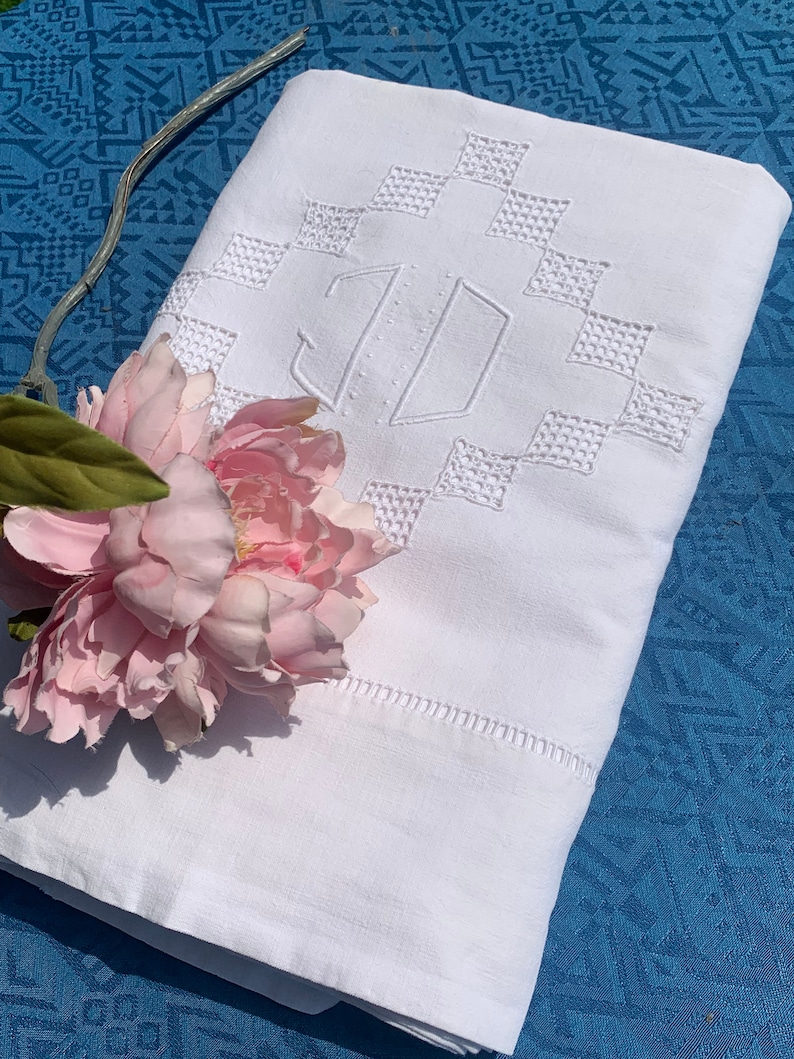 May include: White linen sheet with embroidered geometric design and monogram. A pink peony and a silver decorative element are placed on the sheet. The sheet is folded on a blue patterned surface.