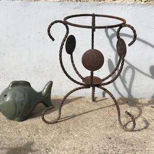 Può includere: Un portavasi in metallo arrugginito con un piano circolare e tre gambe. Il portavasi è realizzato in metallo attorcigliato e presenta un design decorativo.