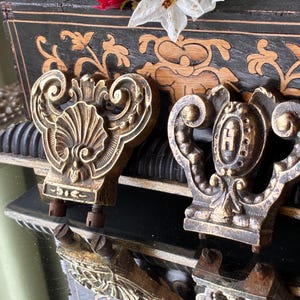 May include: Two antique bronze-colored furniture pulls with ornate designs. One features a shell motif, the other an oval with the letter "H". Both have screw attachments. The background includes a decorative wooden surface.