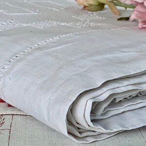 May include: A folded, off-white linen textile with embroidered geometric patterns and a decorative border. The fabric is soft and appears to be vintage, with a slightly textured surface. A pink flower is visible in the background.