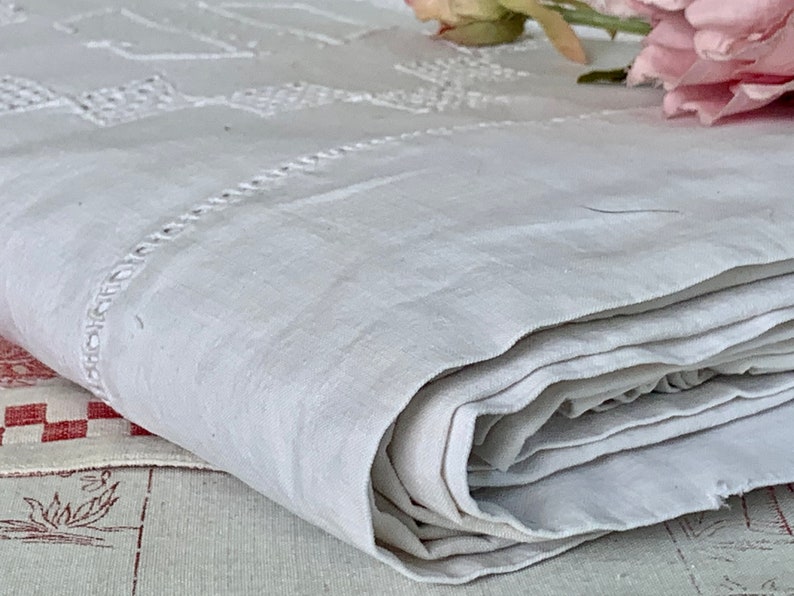 May include: A folded, off-white linen textile with embroidered geometric patterns and a decorative border. The fabric is soft and appears to be vintage, with a slightly textured surface. A pink flower is visible in the background.