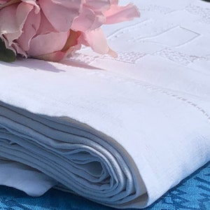 May include: A folded white linen tablecloth with embroidered geometric patterns and a delicate stitched border. A pink flower rests on the corner of the folded cloth. The tablecloth is on a blue surface.