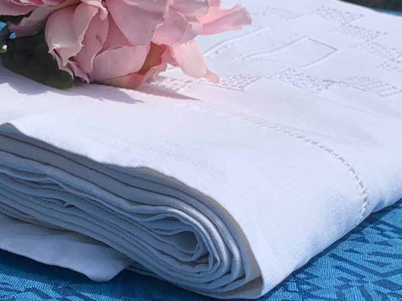 May include: A folded white linen tablecloth with embroidered geometric patterns and a delicate stitched border. A pink flower rests on the corner of the folded cloth. The tablecloth is on a blue surface.