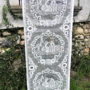 以下が含まれることがあります： 白いレースのテーブルランナー。花輪と建物の詳細なイラストが繰り返されるパターンです。ランナーは屋外に吊り下げられており、背景には石壁と緑があります。レースは繊細でオープンな織り方をしています。