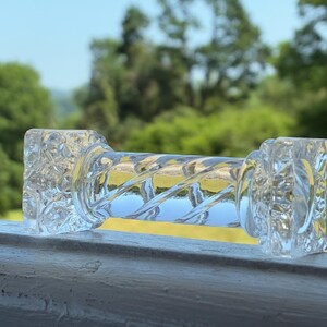 May include: A clear glass dumbbell-shaped object with a textured, square-shaped end and a twisted, cylindrical center. The object is resting on a white surface, with a blurred green background of trees and sky.