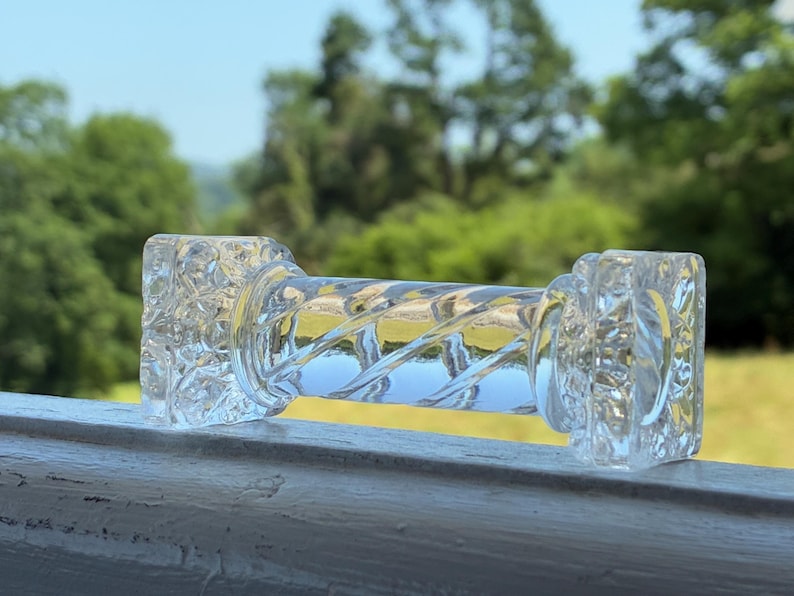 May include: A clear glass dumbbell-shaped object with a textured, square-shaped end and a twisted, cylindrical center. The object is resting on a white surface, with a blurred green background of trees and sky.