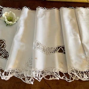 Könnte beinhalten: Ein weißer Tischläufer aus Spitze mit einem gewellten Rand und zarten Blumendesigns. Der Läufer ist in drei Teile gefaltet und hat eine kleine weiße Blume obenauf.