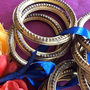 May include: A collection of antique gold-colored metal rings with a textured beaded edge, tied together with a blue satin ribbon. The rings are stacked on a purple surface, with colorful flowers in the background.