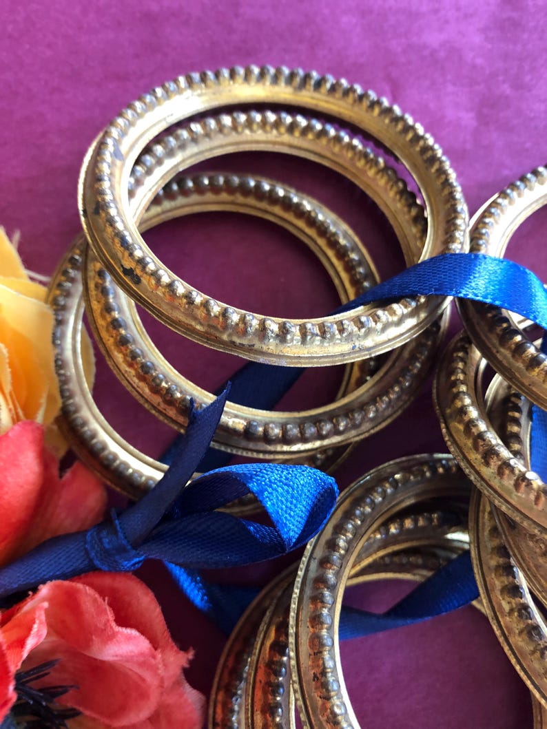 May include: A collection of antique gold-colored metal rings with a textured beaded edge, tied together with a blue satin ribbon. The rings are stacked on a purple surface, with colorful flowers in the background.