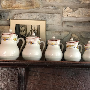 Puede incluir: Cinco jarras de cerámica blanca con diseños florales azules y amarillos y ribete rojo. Las jarras están dispuestas en una fila en un estante de madera.