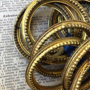 May include: A collection of antique gold-colored oval frames with ornate beaded and textured detailing. The frames are stacked and slightly overlapping, with a blue ribbon visible. The background includes aged paper with French text.