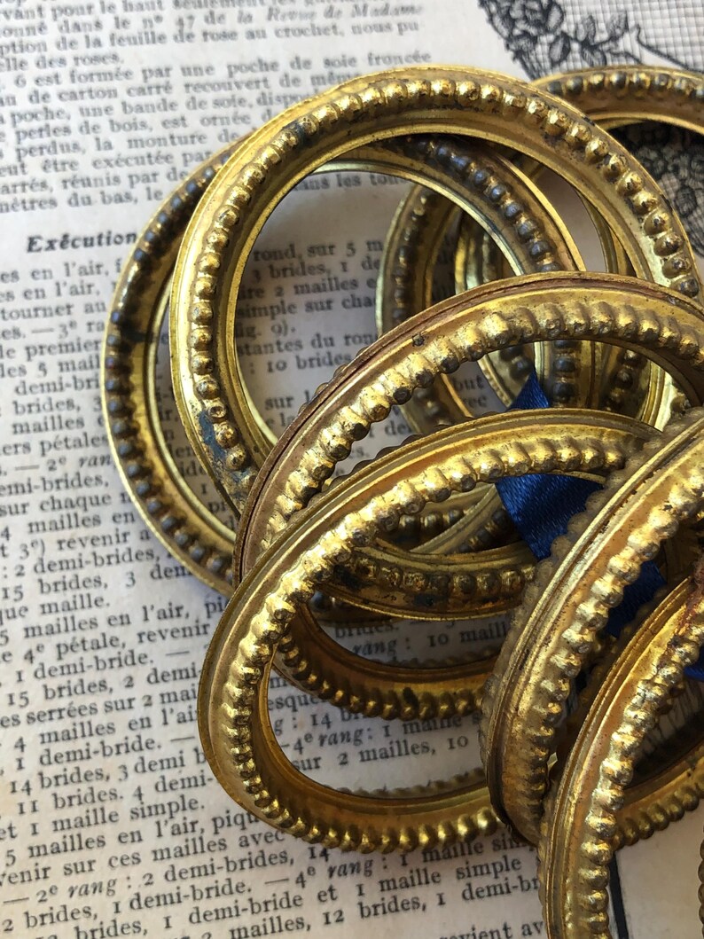 May include: A collection of antique gold-colored oval frames with ornate beaded and textured detailing. The frames are stacked and slightly overlapping, with a blue ribbon visible. The background includes aged paper with French text.